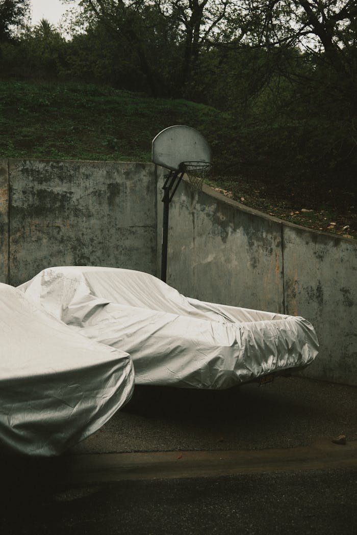 Covered cars beneath a basketball hoop in a moody, outdoor setting with concrete architecture.