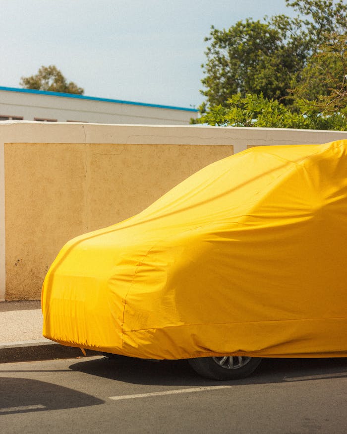 Car covered in bright yellow fabric parked on a street beside a beige wall.